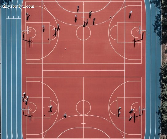 An aerial view of a large, vibrant sports complex featuring a red basketball court with multiple hoops and a surrounding blue running track. Several people are scattered across the area, engaged in various activities.