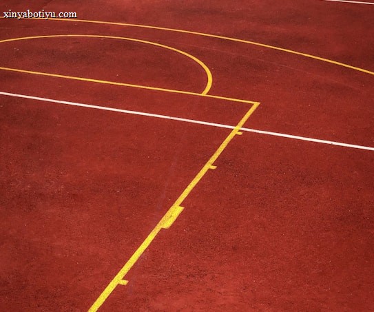 A sports court with a reddish-brown surface and bright yellow and white lines. The lines form geometric shapes, including rectangles and circles, suggesting markings for games like basketball or netball.